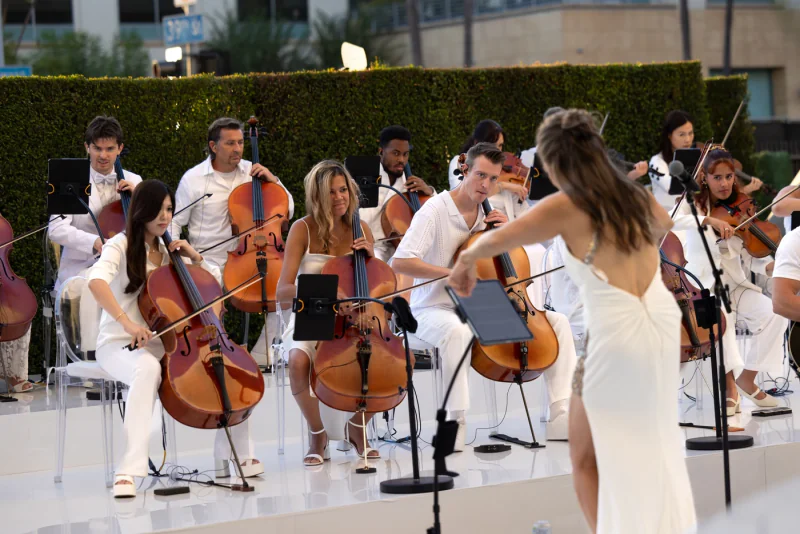 A live orchestra with cellists and conductor in a sunlit outdoor setting